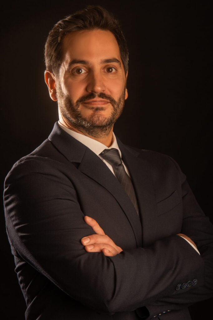 Un homme en costume se tient avec confiance, souriant légèrement.